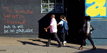 Two adults and a school child walk down a street, past graffiti which says, 'The revolution will not be televised, but it will be on YouTube.'